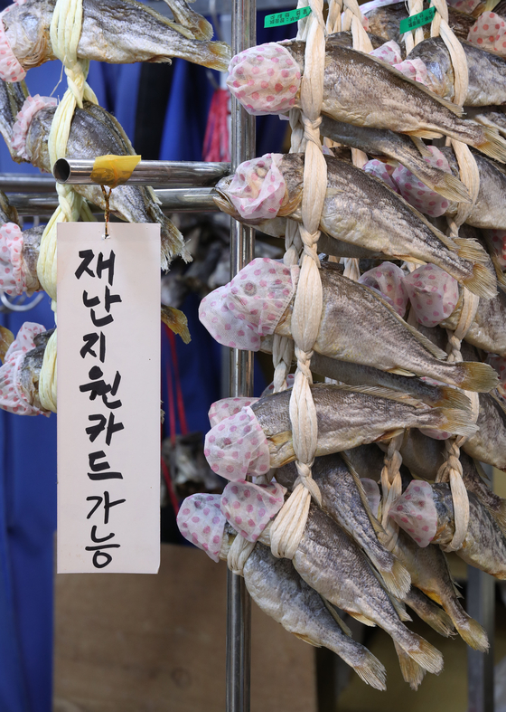 A shop displays dried fish with microphone covers over the heads at a local market in central Seoul on Friday next to a sign that says the store accepts the government’s emergency relief grant. Since relief grants were released on May 13, spending at local stores has gone up, contributing to a sales boost for small businesses. [YONHAP]