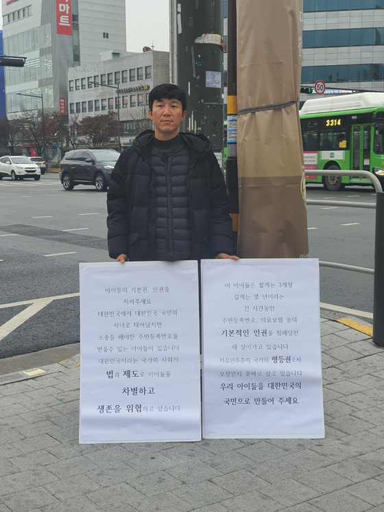 Kim Ji-hwan holding a one-person protest in the streets of Seoul with cardboard signs that read, “Please protect the rights of children [...] These children are forced to live for at least three months to as long as years without a social security number or health insurance.” [KIM JI-HWAN]