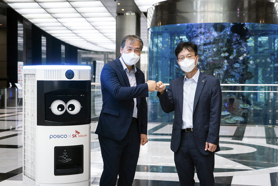 Posco's head of Process & Engineering R&D Center Kim Ki-soo, left, and SK Telecom's head of the Smart Factory division, Choi Nak-hoon, pose for photos after signing a business agreement on Friday. Standing to the left of Kim is Keemi, SK Telecom's automated cleaning and disinfecting machine. [SK TELECOM]