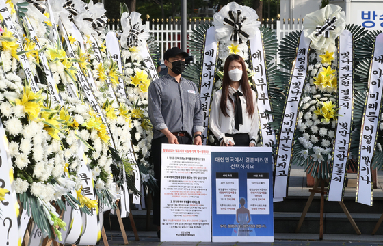 Surrounded by standing wreathes that were sent by dozens of frustrated couples across the nation, a couple holds a sign demanding the easing of government social distancing restrictions on weddings at a protest sponsored by the Federation of Korean Newlyweds on Thursday morning. [NEWS1]