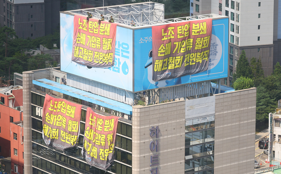 Three truck drivers in the Cargo Truckers Solidarity sit on top of a billboard installed at HiteJinro’s headquarters in Gangnam District, southern Seoul. Around 100 unionized workers occupied the building lobby and rooftop on Tuesday. [YONHAP]