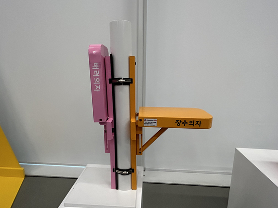 The orange chair on the right is a prototype “Longevity Chair,” a foldable seat installed on columns at crosswalks for the elderly. The pink one on the left, the “Caring Chair,” is for anyone who needs it, including children or pregnant women. [SHIN MIN-HEE]