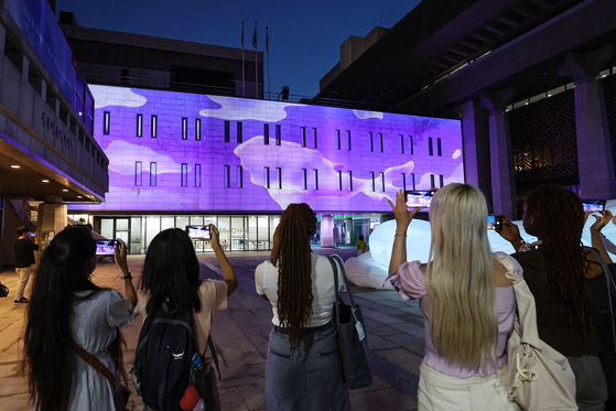 Tourists take pictures as the outer wall of the Sejong Center for the Performing Arts in central Seoul is shone with purple lighting on June 13, as part of the ″2023 BTS Festa″ held in celebration of K-pop boy band BTS's 10th anniversary of debut that fell that day. [YONHAP]