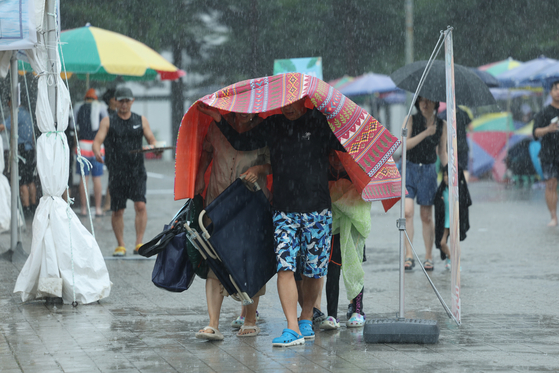 A family uses a picnic mat to take cover from a downpour at Ttukseom Resort near the Han River in Gwangjin District, western Seoul, on Sunday morning. The Korea Meteorological Administration said heavy showers will fall over the country all through the week as the summer monsoon front stalls over the Korean Peninsula. [YONHAP]