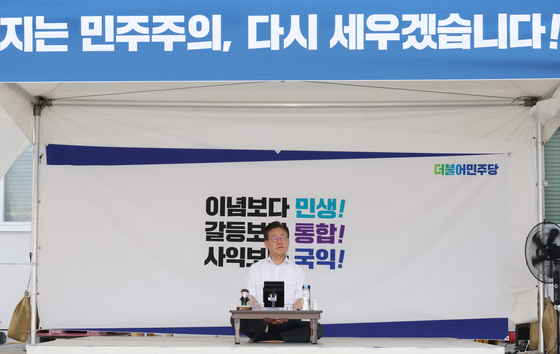 Democratic Party leader Lee Jae-myung sits under a white canopy in front of the National Assembly in Yeouido, western Seoul, on Thursday, the first day of his indefinite hunger strike against the Yoon Suk Yeol administration. The banner above him vows to “rebuild” Korea's “crumbling democracy.” [YONHAP]