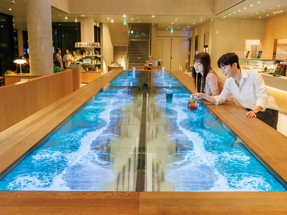 People look at the 55-inch transparent organic light-emitting diode (OLED) installation at Starbucks’ Yeosu Dolsan DT branch in South Jeolla. LG Display and Starbucks Korea collaborated to install the OLED table in the store, which shows images of rolling waves on the beach. The display has sensors that change the waves based on the coffee mugs sitting on the table. [LG DISPLAY]