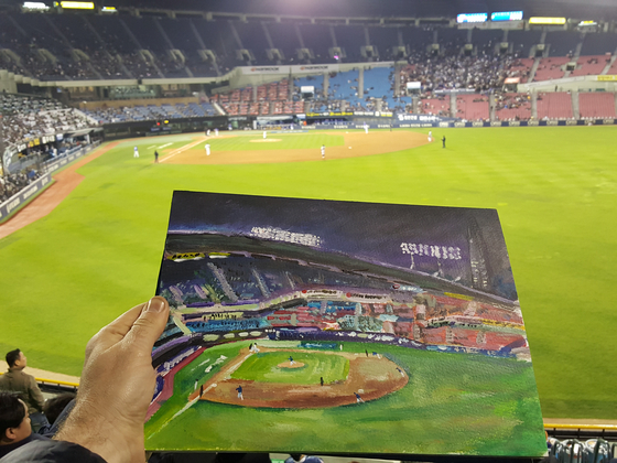 Andy Brown holds a painting of the field at Jamsil Baseball Stadium in front of the same view from the stands at Jamsil Baseball Stadium in southern Seoul.  [ANDY BROWN]
