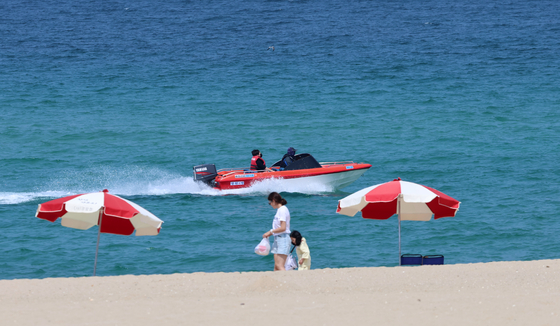 A motor boat cruises along Gyeongpo Beach in Gangneung, Gangwon, on Tuesday, when daytime temperatures reached 34.4 degrees Celsius (93.9 degrees Fahrenheit). Two people stroll on the beach past two parasols planted in the sand.[YONHAP]