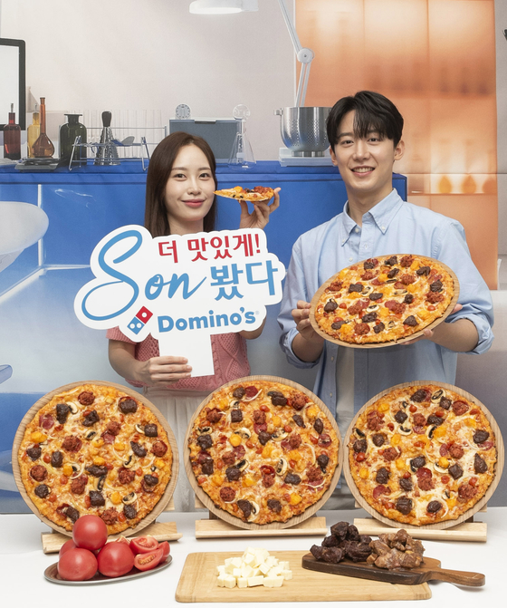 Models hold up Domino Pizza's upcoming menu item, K-Rib & Cheese Pizza, set to be released on Friday.  The Pizza features ″Sonny cube cheese,″ inspired by Son Heungmin, the brand's ambassador, and his signature pose. [DOMINOS PIZZA]