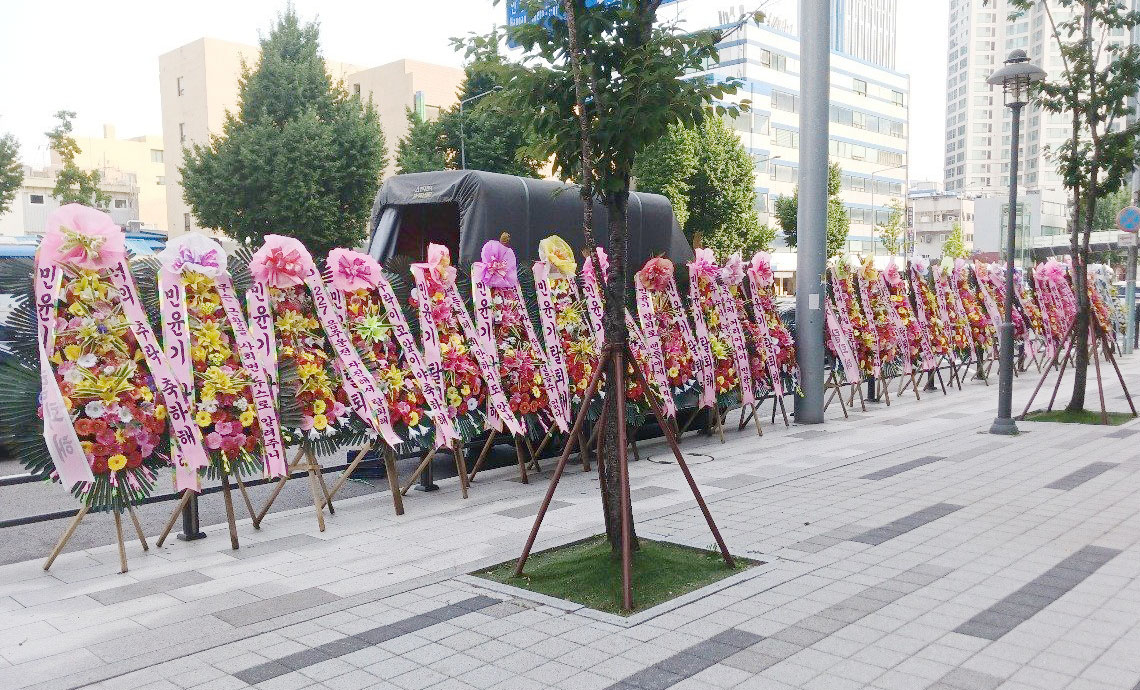 Flower wreaths displaying messages demanding BTS's Suga to leave the band are positioned in front of HYBE's headquarters in Yongsan District, central Seoul, on Tuesday. [SCREEN CAPTURE]